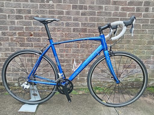 A blue road bicycle with drop handlebars, resting against a brick wall.